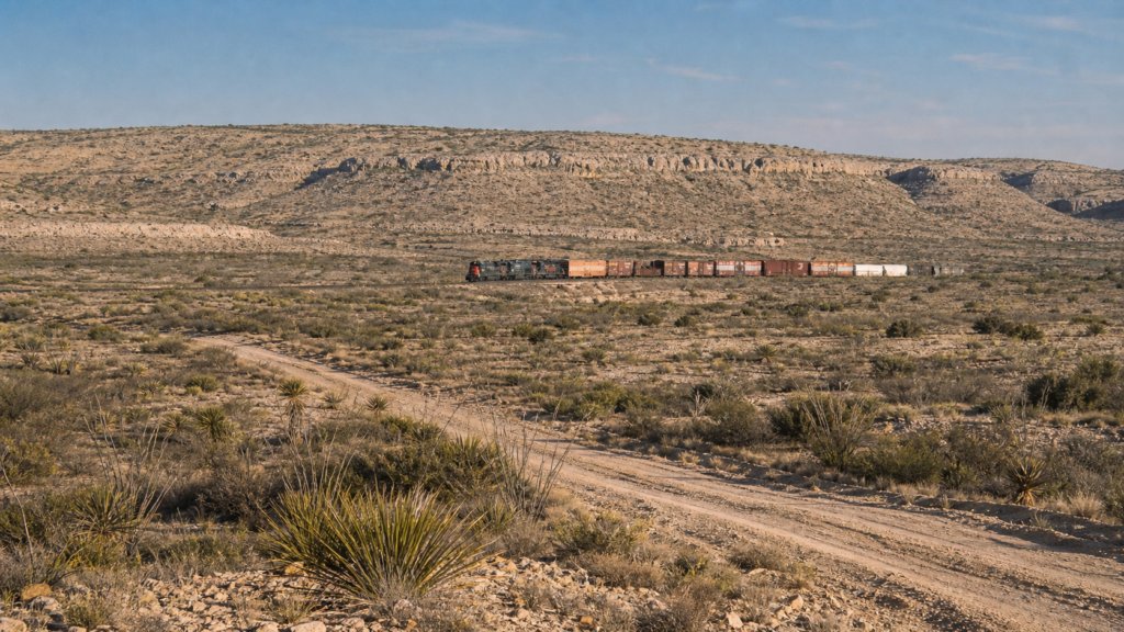 A long freight train led by red-and-black locomotives curves through rugged, dry Terrell County–style West Texas desert beneath limestone cliffs. 